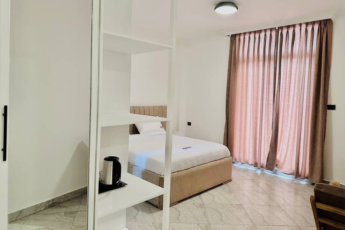 Ein Schlafzimmer mit Bett, Schrank, Ablagefläche mit Wasserkocher und Fenster mit Vorhängen.