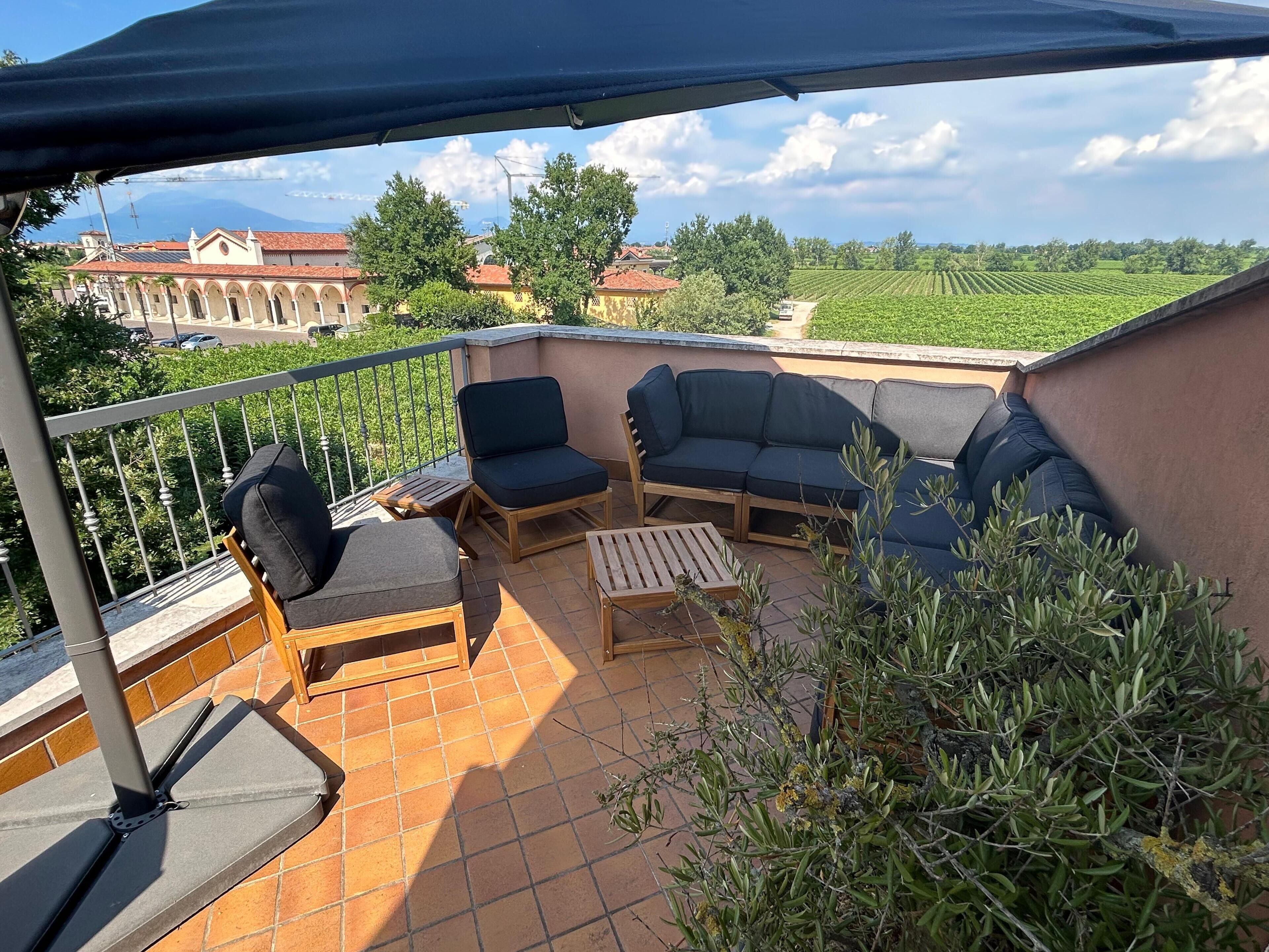 A terrace with outdoor furniture, a view of vineyards, and a building with red-tiled roofs in the background.