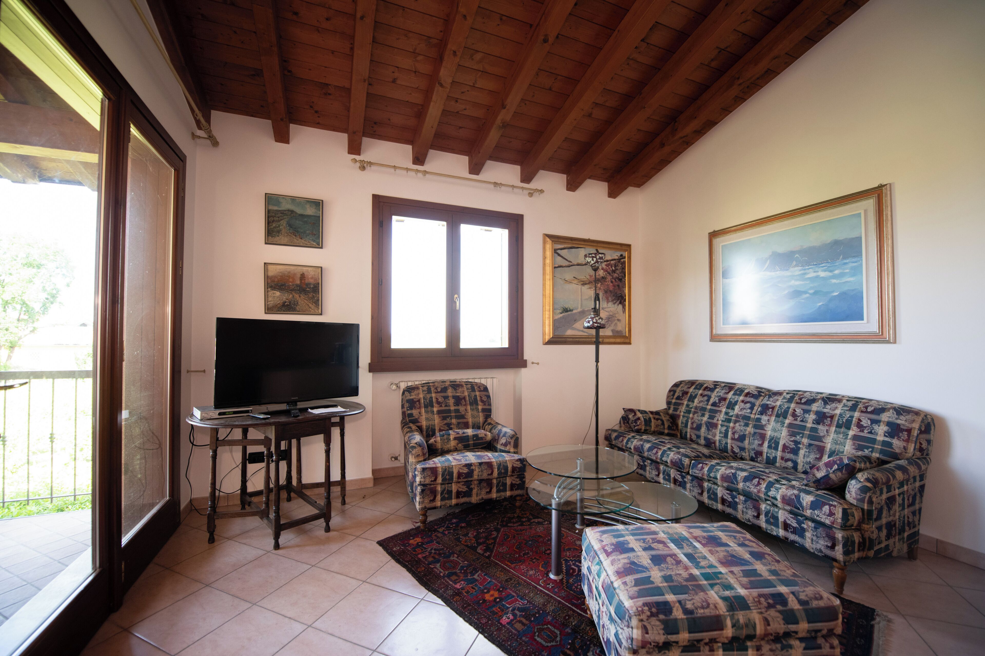 A living room with a television, two patterned armchairs, a sofa, a glass coffee table, and a large window.