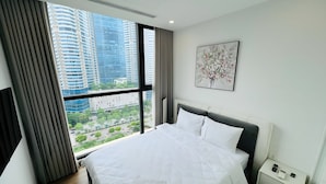 A modern hotel room with a large bed, a bedside table with a phone and charger, a window with a city view, and a painting on the wall.