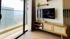 A modern living room with a large flat-screen TV mounted on the wall, wooden shelves, and a balcony with a city view.