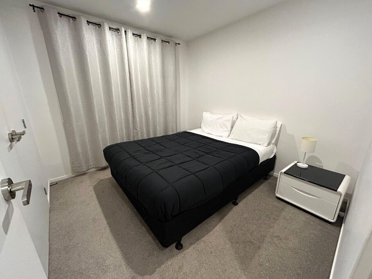 A bedroom with a black bed, white curtains, a white bedside table with a lamp, and a door.