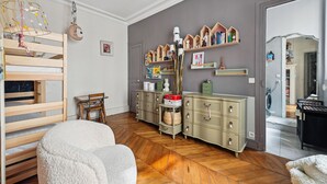 A child's room with a bunk bed, a dresser, shelves with books and toys, and a door leading to another room.