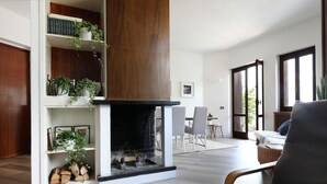 Una sala de estar moderna con chimenea, estanterías de madera con plantas, un comedor y un ventanal grande.