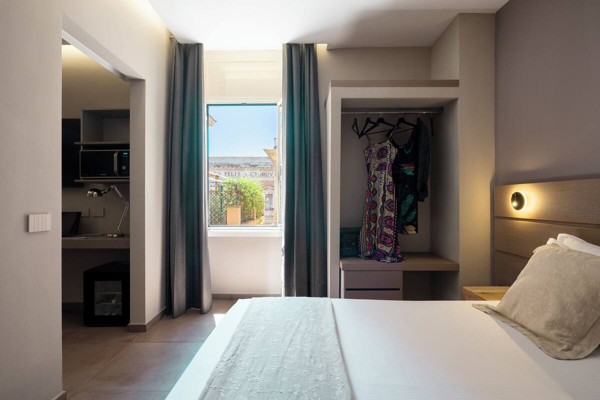 A hotel room with a bed, a desk with a computer, a wardrobe, and a view of the outside.