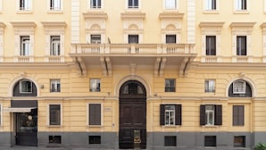 Un bâtiment jaune avec une porte noire au centre, de nombreuses fenêtres avec des volets et un balcon doté de balustrades.