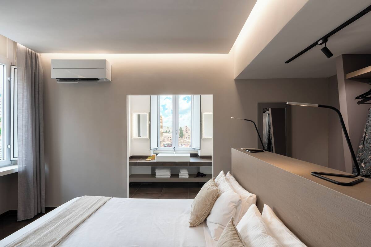A modern hotel room with a large bed, a built-in desk, a mirror, and a view of the city through a large window.