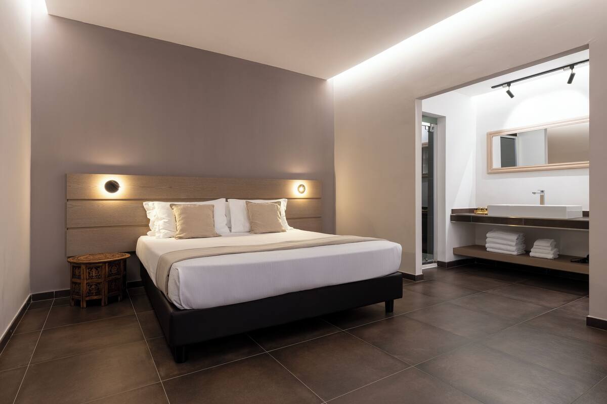 A modern bedroom with a large bed, bedside tables, a built-in bench, and a bathroom with a vanity and mirror.