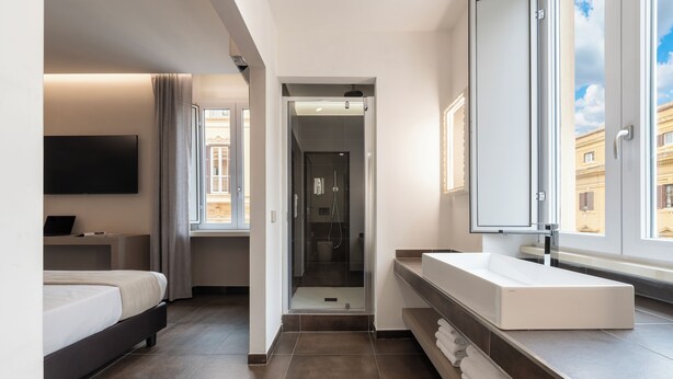 Une chambre d’hôtel moderne dotée d’un grand lit, d’une télévision à écran plat, d’une salle de bain avec un grand miroir et offrant une vue sur la ville par la fenêtre.