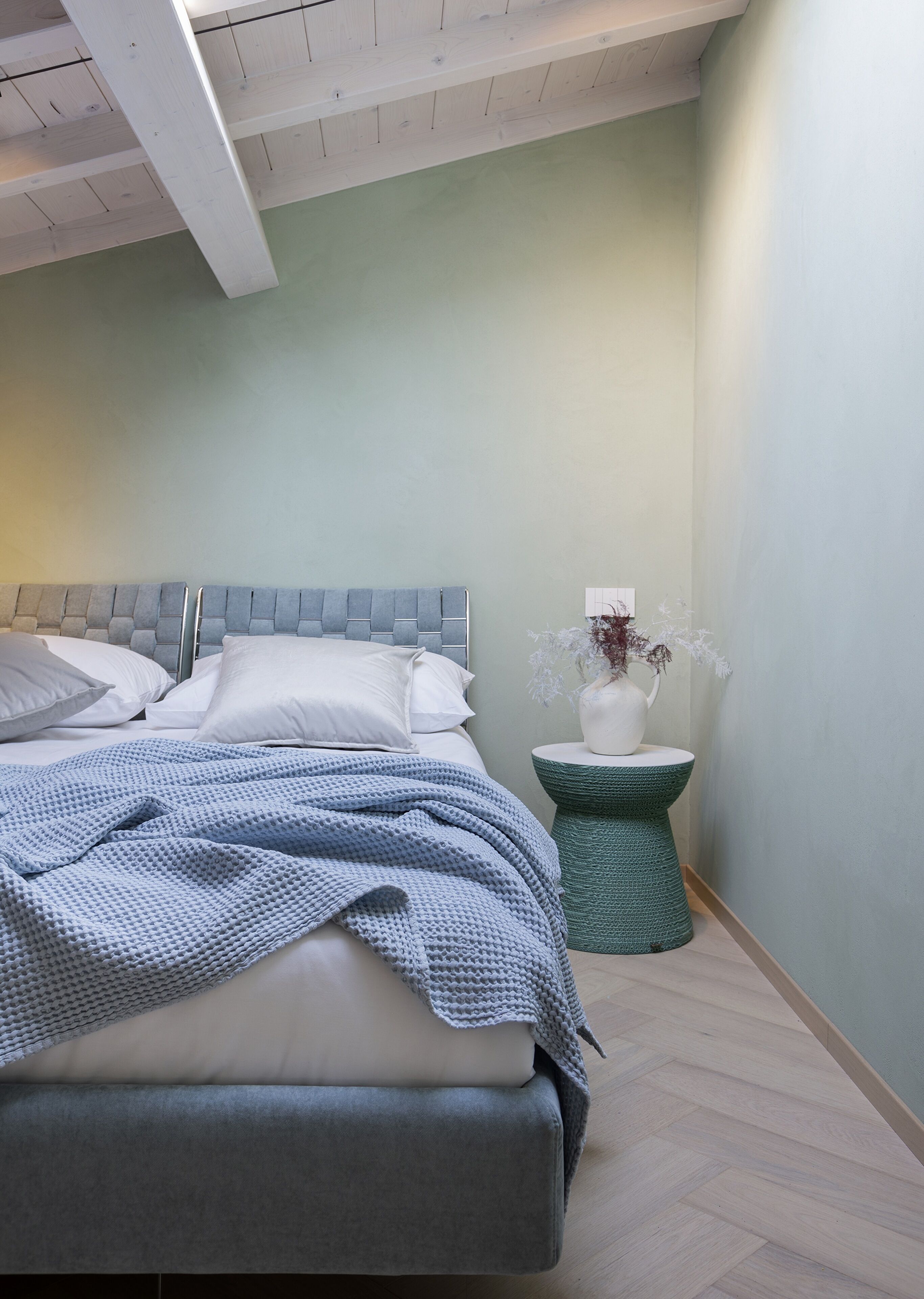 Un dormitorio con una cama, almohadas, una colcha azul, una mesita de noche con un florero y un taburete verde.