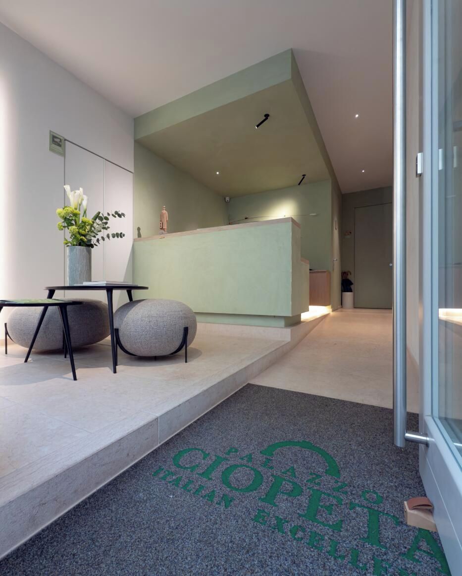 Una moderna área de recepción con diseño minimalista, que incluye un mostrador de recepción, una zona de estar con taburetes redondos y un jarrón decorativo con flores.