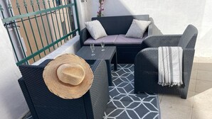 Un balcón con un sofá negro tejido, un sillón a juego, una mesita con dos copas de vino y un sombrero tejido.
