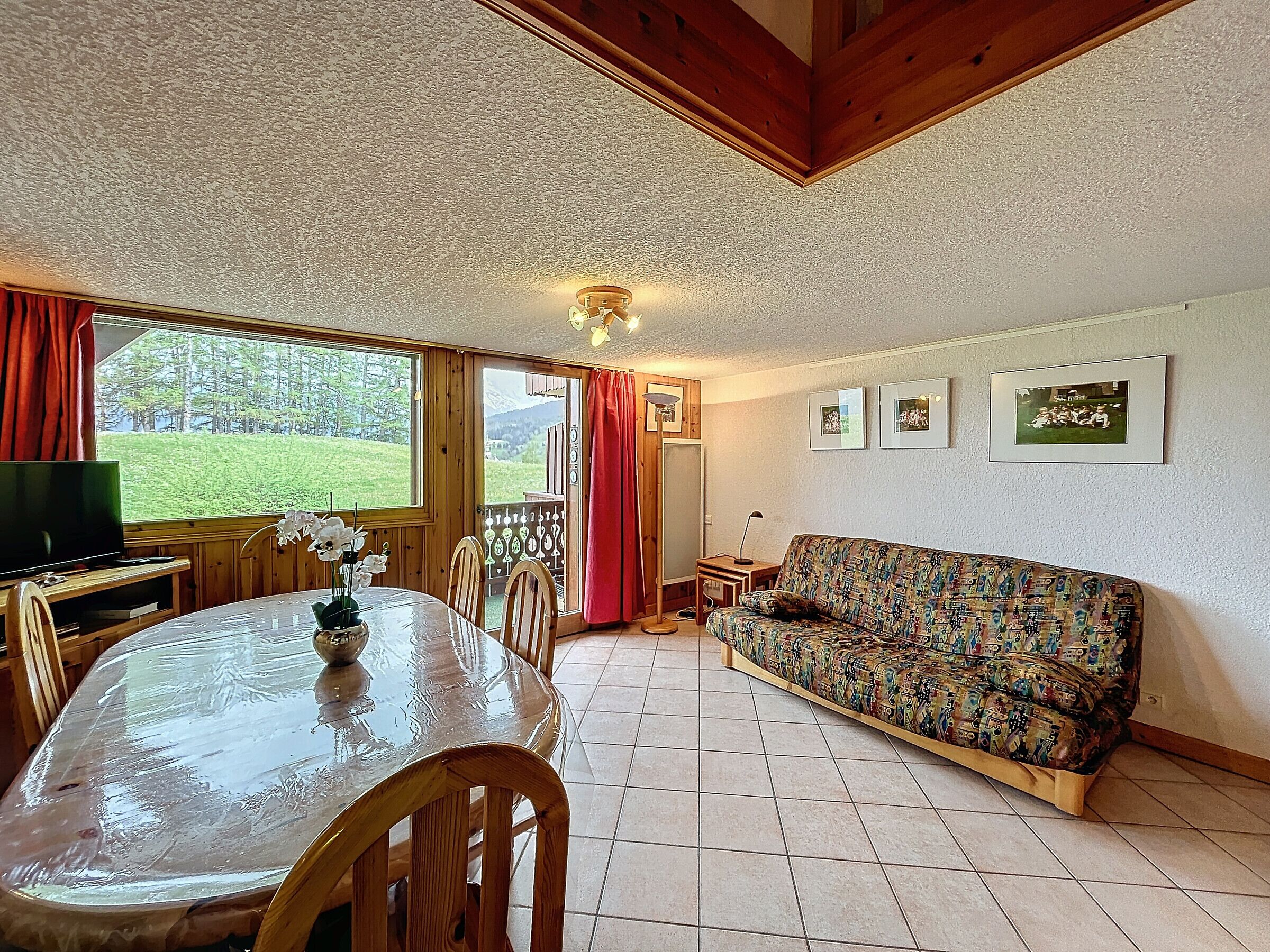 A cozy living room with a wooden ceiling, a sofa, a dining table with chairs, and a TV.