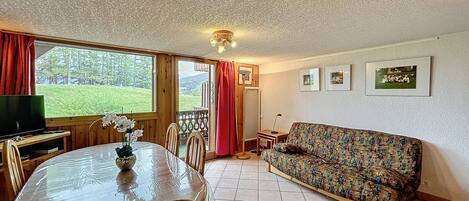 A cozy living room with a wooden ceiling, a sofa, a dining table with chairs, and a TV.