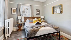 Une chambre à coucher avec un lit, des tables de chevet, un radiateur, une fenêtre avec des rideaux et un miroir au mur.