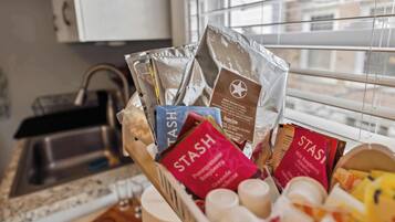 A kitchen counter with a basket containing various coffee pods and a window with blinds.