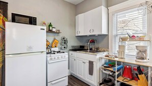 Une cuisine avec des meubles blancs, une cuisinière, un four à micro-ondes et un évier. On y trouve des étagères remplies de divers ustensiles de cuisine et une fenêtre avec des stores.