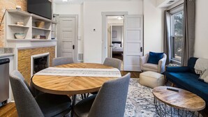 A modern living room with a wooden dining table, grey upholstered chairs, a blue sofa, and a TV mounted on a brick wall. - Victorian Estates | Sun Soaked 1BR | Close to CCF (Cleveland)