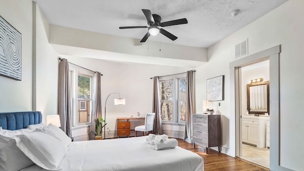 A bedroom with a bed, a desk, a chair, a dresser, and a bathroom visible through an open door. - Victorian Estates | Sun Soaked 1BR | Close to CCF (Cleveland)