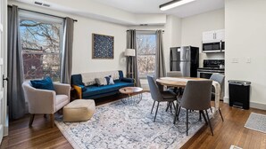 A modern living room with a blue sofa, a dining table with chairs, and a kitchen area with a refrigerator and microwave. - Victorian Estates | Sun Soaked 1BR | Close to CCF (Cleveland)