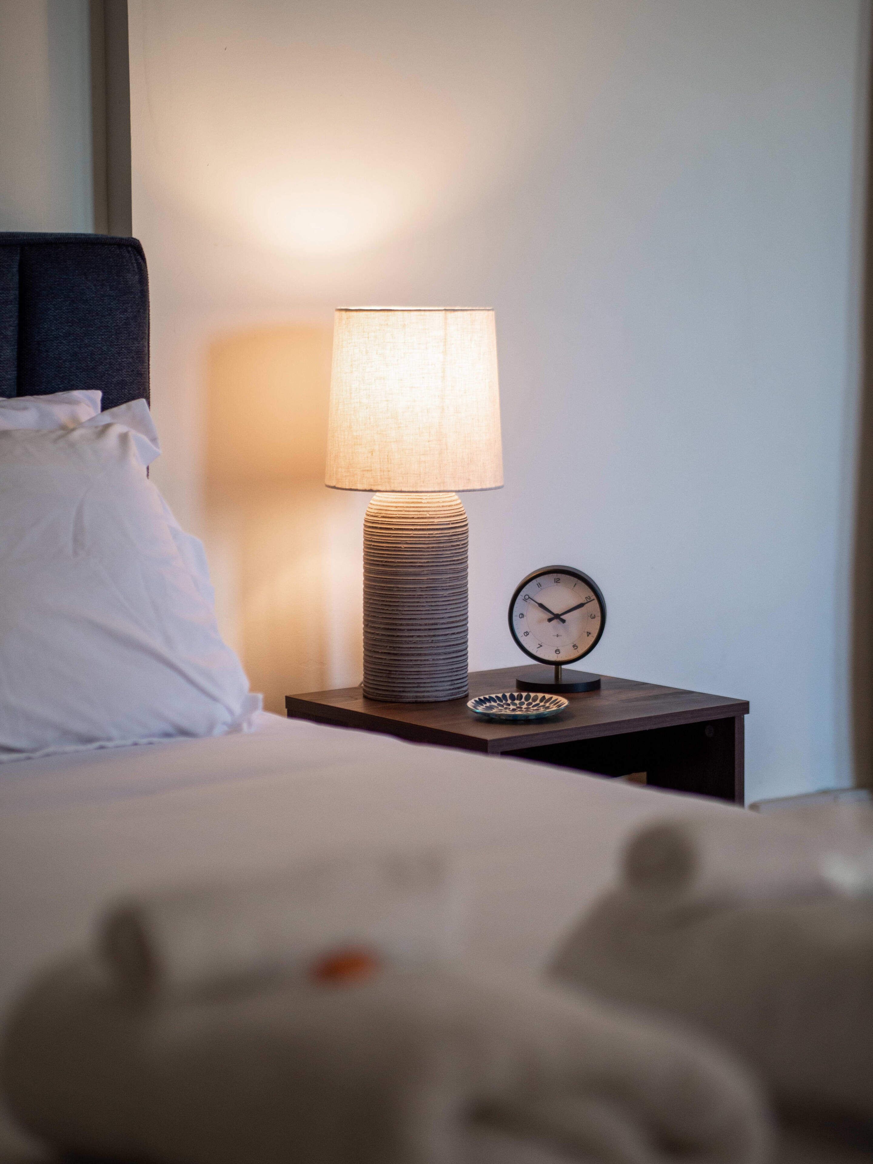 A neatly made bed with white bedding, a bedside lamp with a textured base, a small round clock, and a decorative plate on a wooden nightstand.