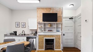 A modern kitchen with a brick accent wall, a flat-screen TV, and a built-in fireplace. - Victorian Estates | Sun Soaked 1BR | Close to CCF (Cleveland)