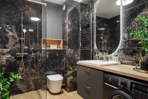 A modern bathroom with a marble wall, a glass-enclosed shower, a toilet, a sink with a mirror, and a washing machine. - Lumina luxury apartment with garden (Łódź)