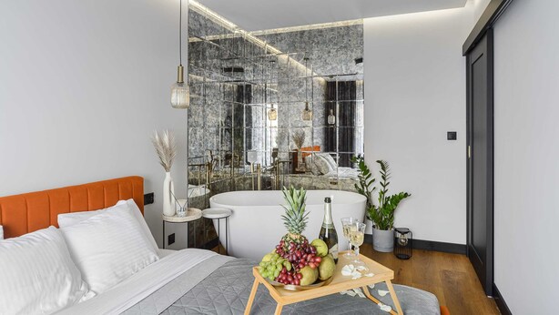 Quarto moderno com cama, mesa de cabeceira com bandeja de frutas, espelho grande e banheira.