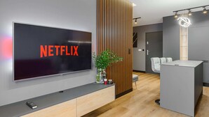 Uma sala de estar moderna com uma grande TV de tela plana exibindo 'NETFLIX', uma parede de ripas de madeira, uma pequena planta, um controle remoto e uma área de estar com um sofá cinza e uma mesinha.