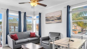 A modern living room with a grey sofa, a wooden coffee table, a metal-framed dining table, and a ceiling fan. - Spectacular Ocean View Corner Apartment (Hollywood)