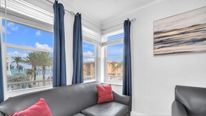 A room with a grey sofa, two red cushions, a painting on the wall, and a view of palm trees and buildings through large windows with blue curtains. - Spectacular Ocean View Corner Apartment (Hollywood)