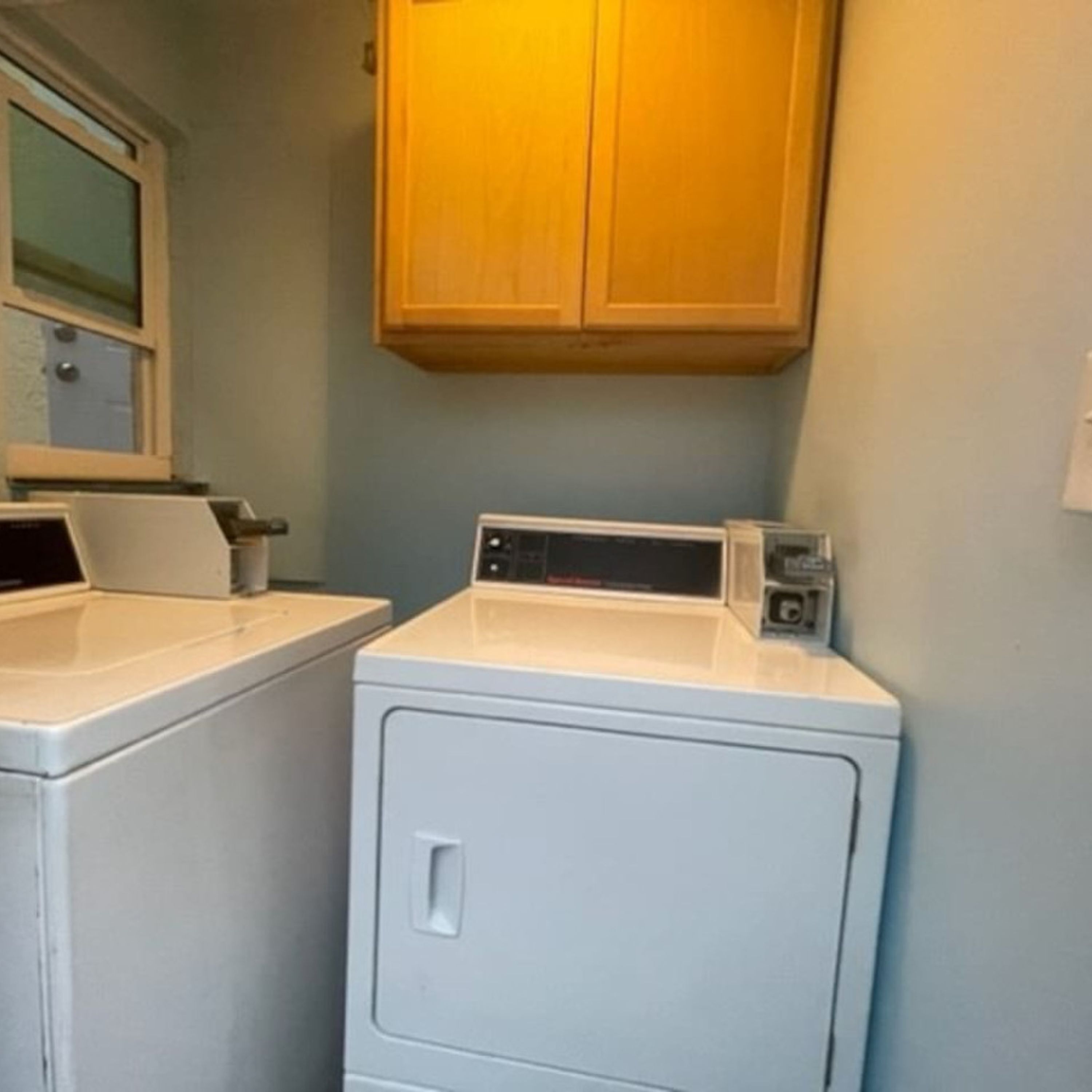 A laundry area with a front-loading washer and dryer, wooden cabinets above, and a window with blinds.
