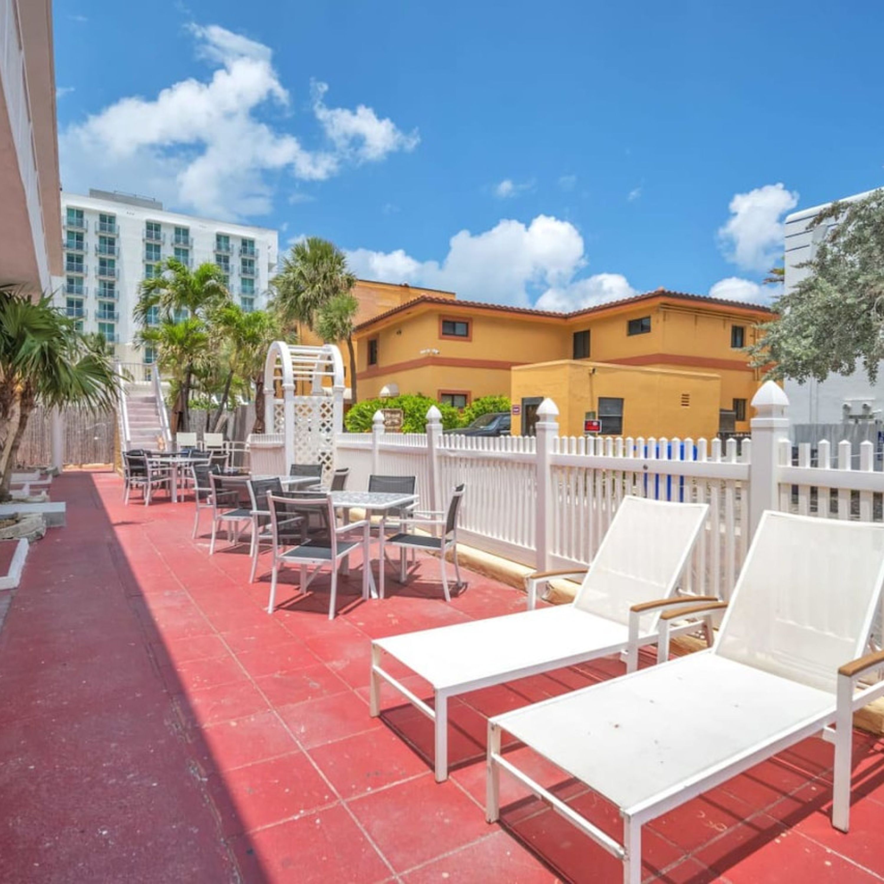 A rooftop patio with white lounge chairs and tables, a white fence, and a yellow building in the background.