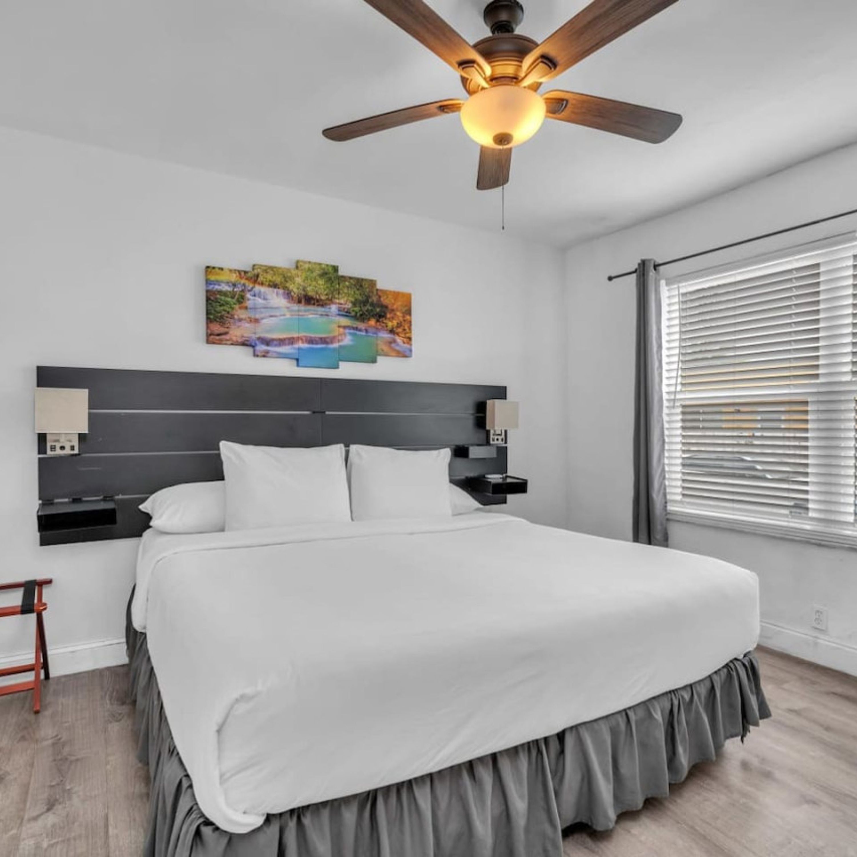 A modern bedroom with a large bed, a ceiling fan, a painting of a waterfall, and a window with blinds.