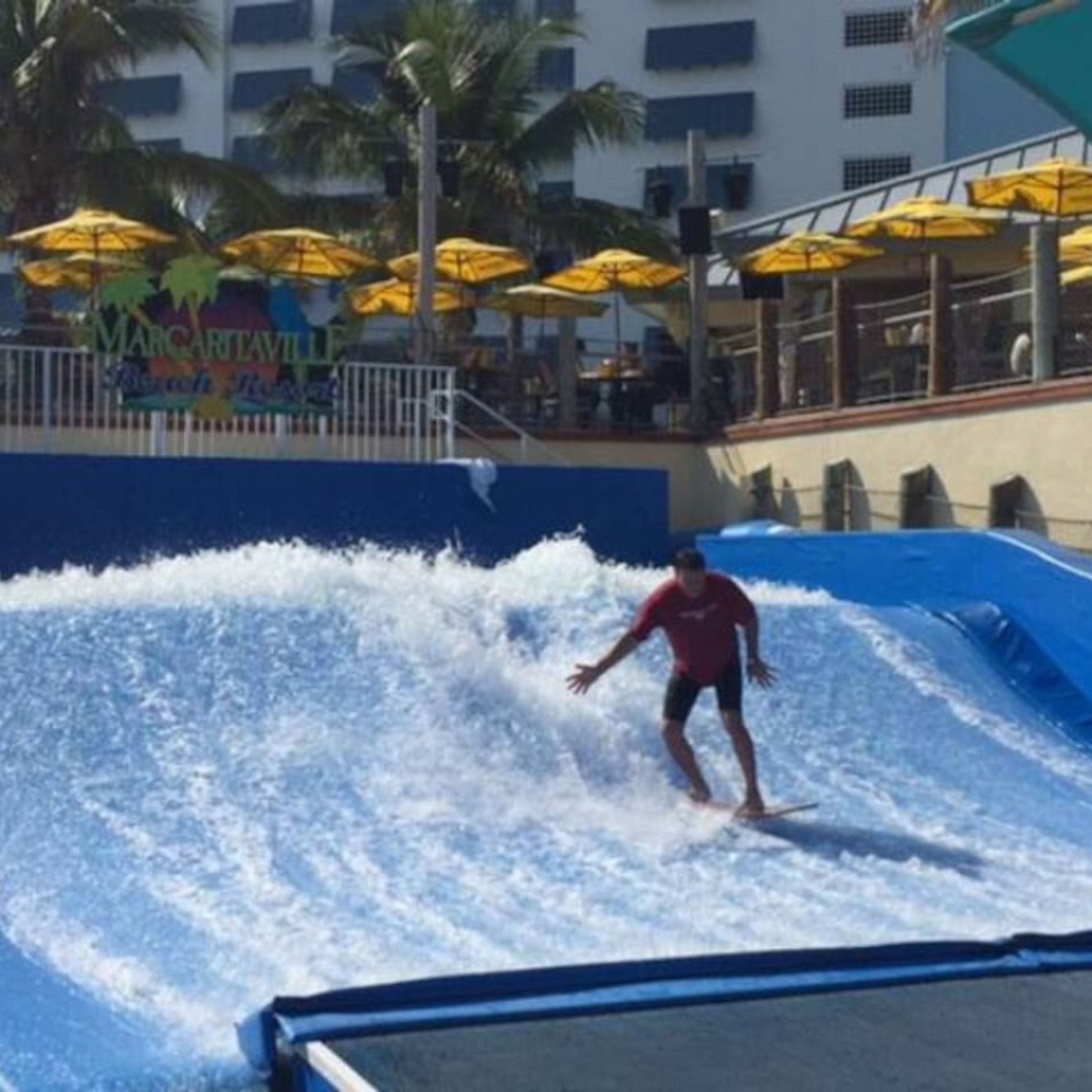 A wave pool with a surfer riding a wave, surrounded by yellow umbrellas and palm trees.