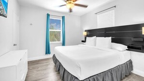 Une chambre à coucher avec un lit, un ventilateur de plafond, une fenêtre avec des rideaux, un téléviseur fixé au mur et une commode blanche.