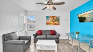 A modern living room with a grey leather sofa, a white coffee table, a dining table with chairs, a ceiling fan, and a painting of a sunset.