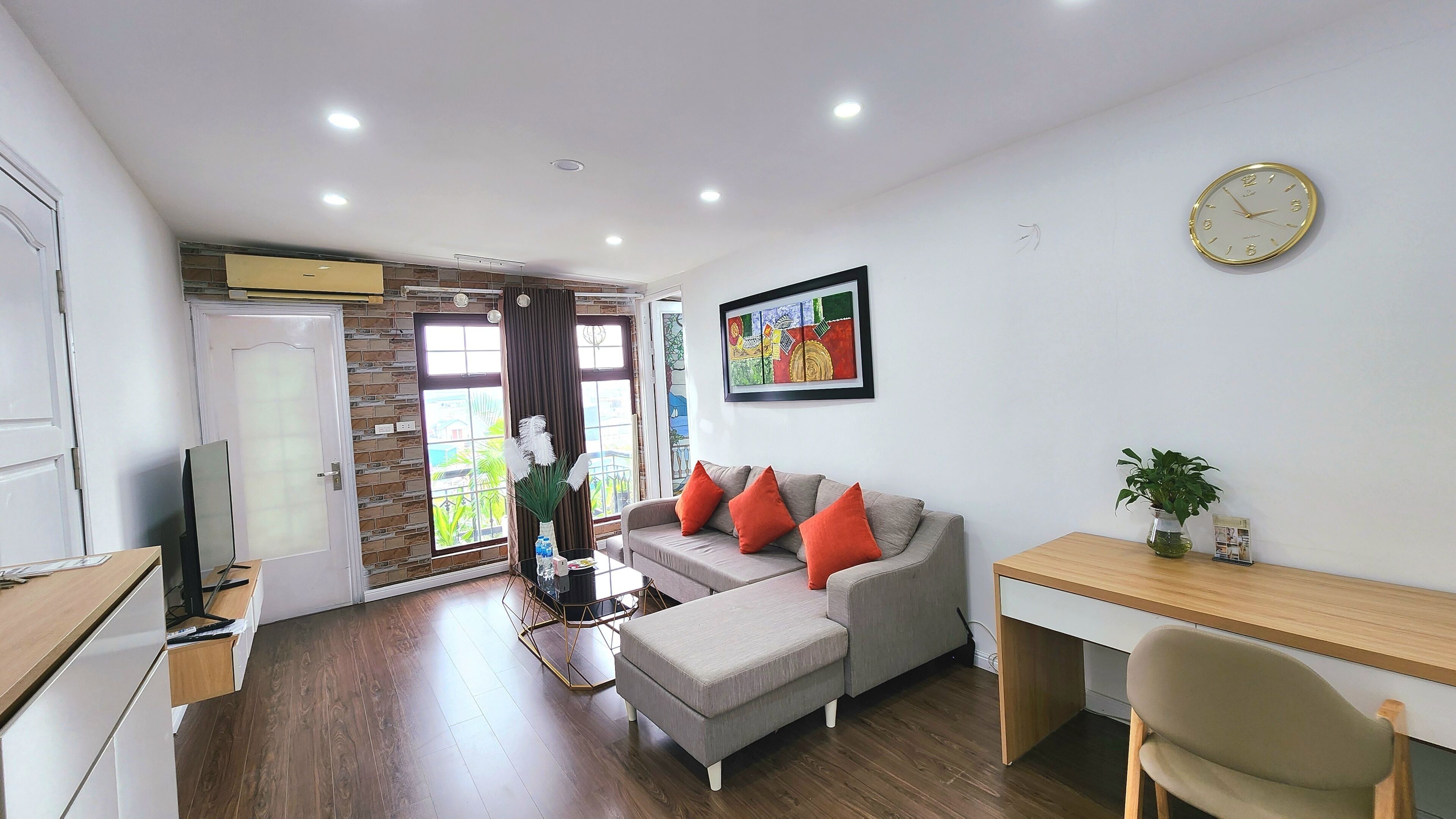 A modern living room with a grey sofa, red cushions, a wooden desk, and a large window with curtains.