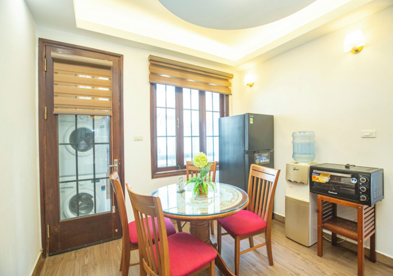 A compact living space with a dining area featuring a round glass table and wooden chairs, a kitchenette with a water dispenser, and a small refrigerator.
