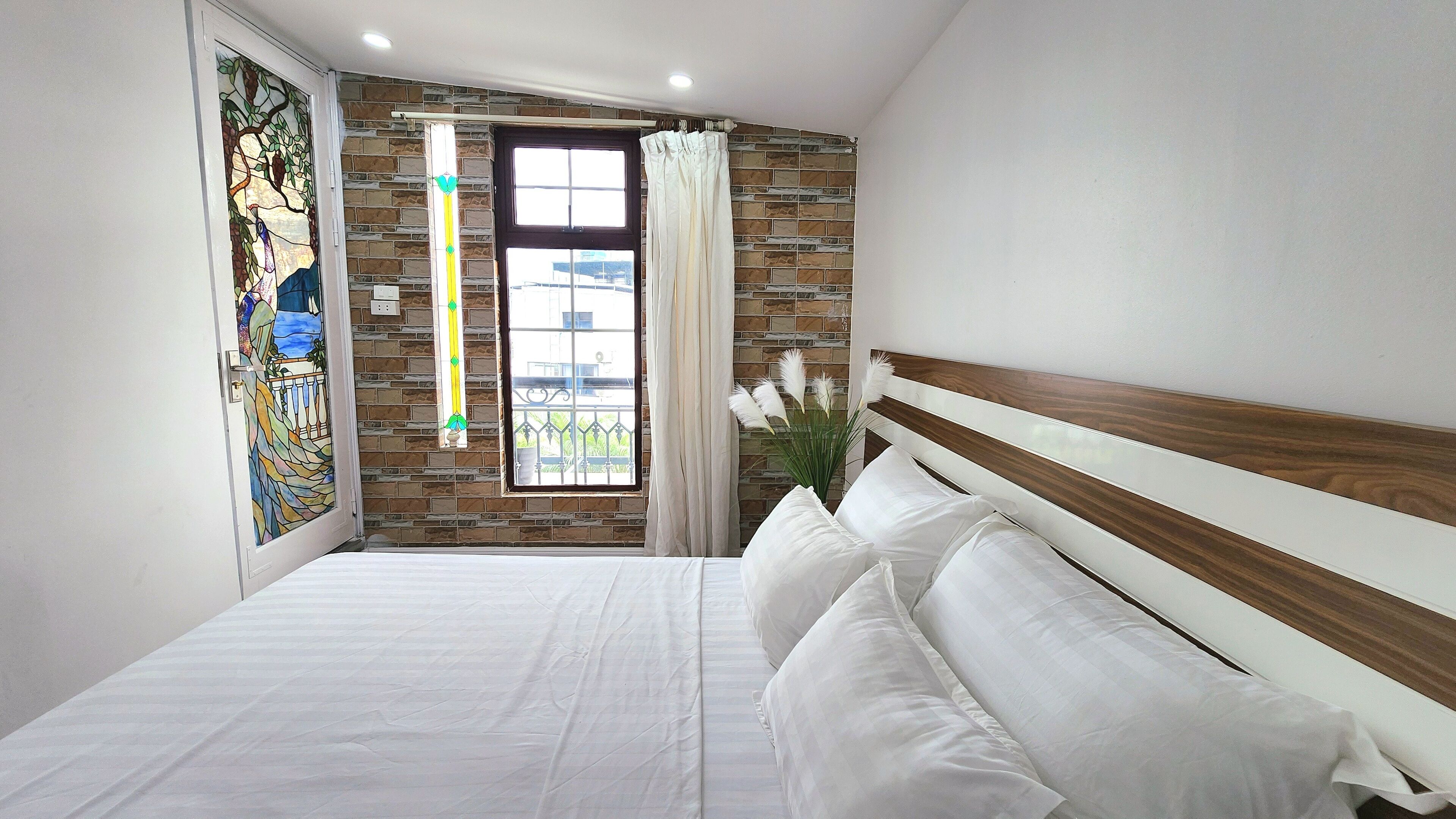 A bedroom with a bed, a window with curtains, a stained glass window, and a brick wall.