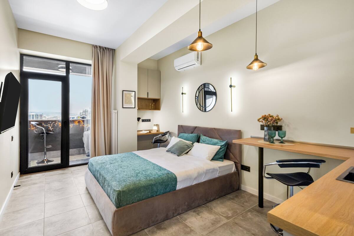A modern hotel room with a bed, desk, and chair, a view through a sliding door, and a ceiling light fixture.