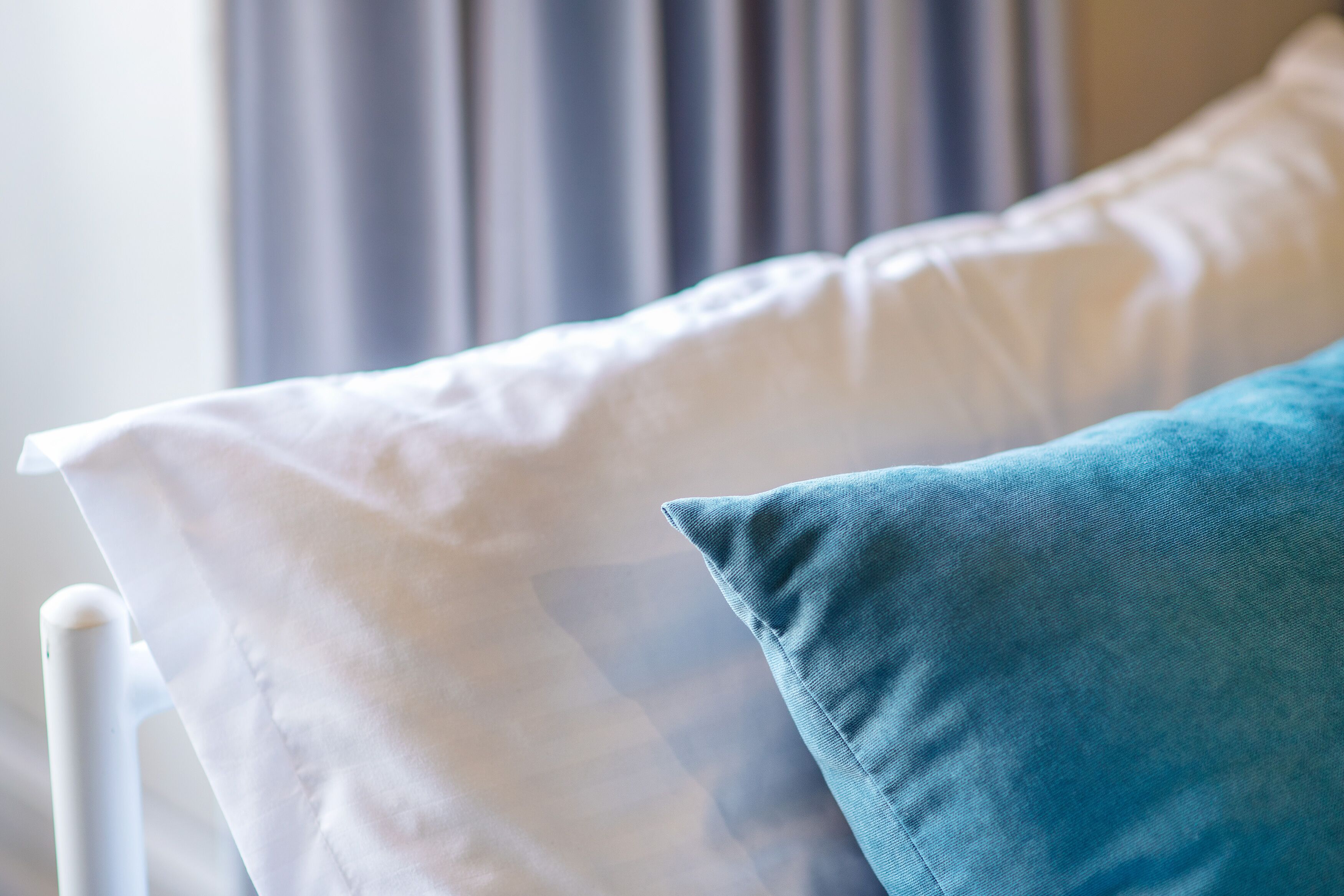 A bed with white and blue pillows, a white headboard, and a window with curtains in the background.