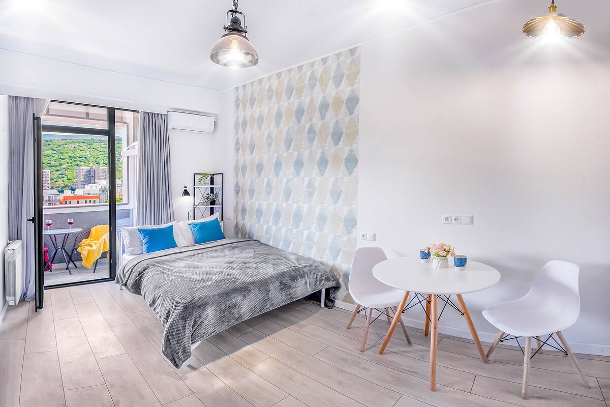 A modern bedroom with a bed, a dining table with chairs, and a view of the city through a glass door.
