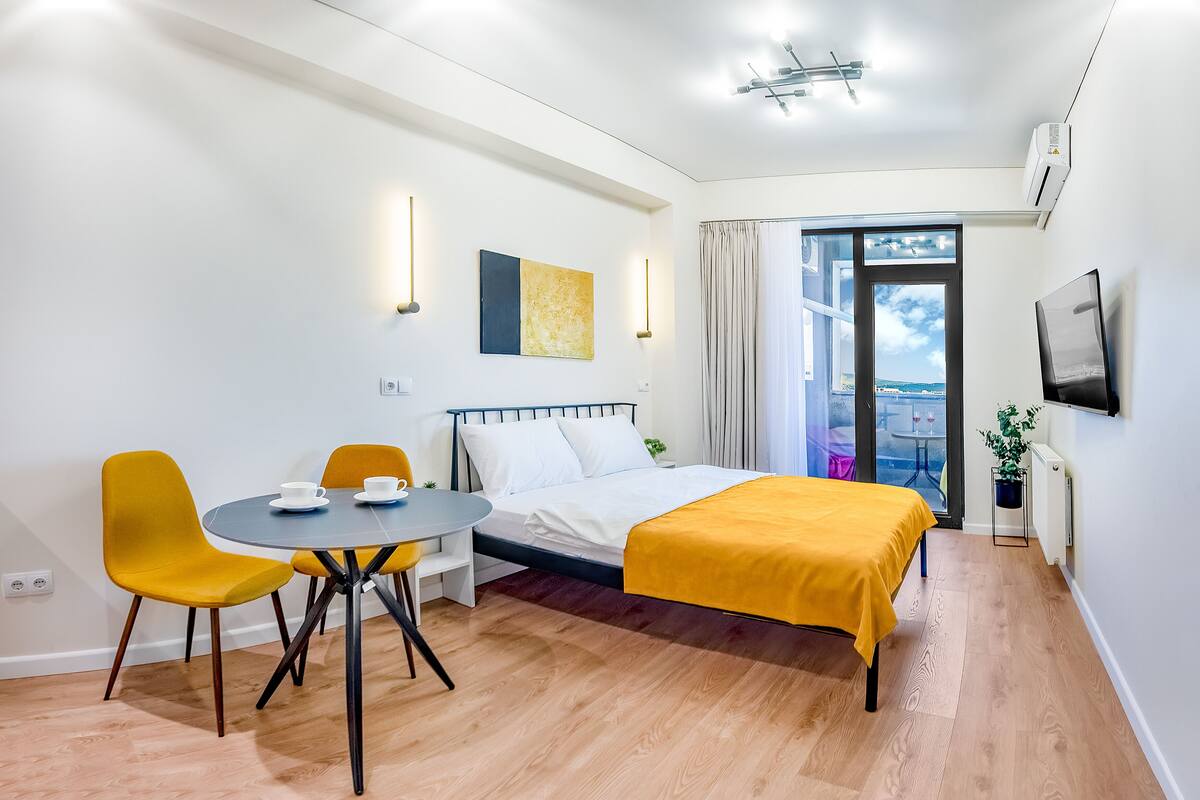 A modern hotel room with a bed, a dining table with chairs, a small balcony, and a TV mounted on the wall.