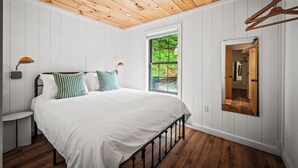 A bedroom with a wooden ceiling, white walls, a large bed with white bedding, a window with a view of greenery, and a mirror on the wall.