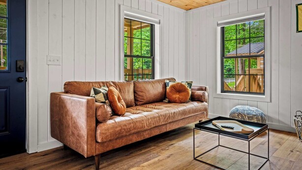 A room with a brown leather sofa, two windows, a wooden ceiling, and a small table with a tray.