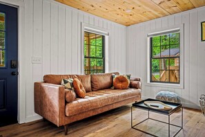 A room with a brown leather sofa, two windows, a wooden ceiling, and a small table with a tray. - Cozy 2BR Cabin in Gatlinburg: Fire Pit & Fun! (Gatlinburg)