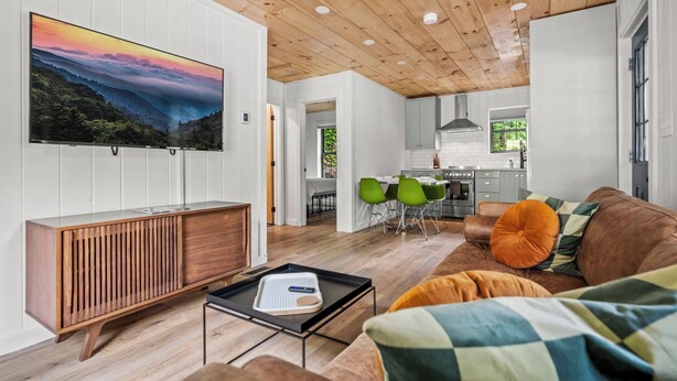 Een moderne woonkamer met een houten plafond, een grote televisie aan de wand, een bruine leren bank en een eethoek op de achtergrond.