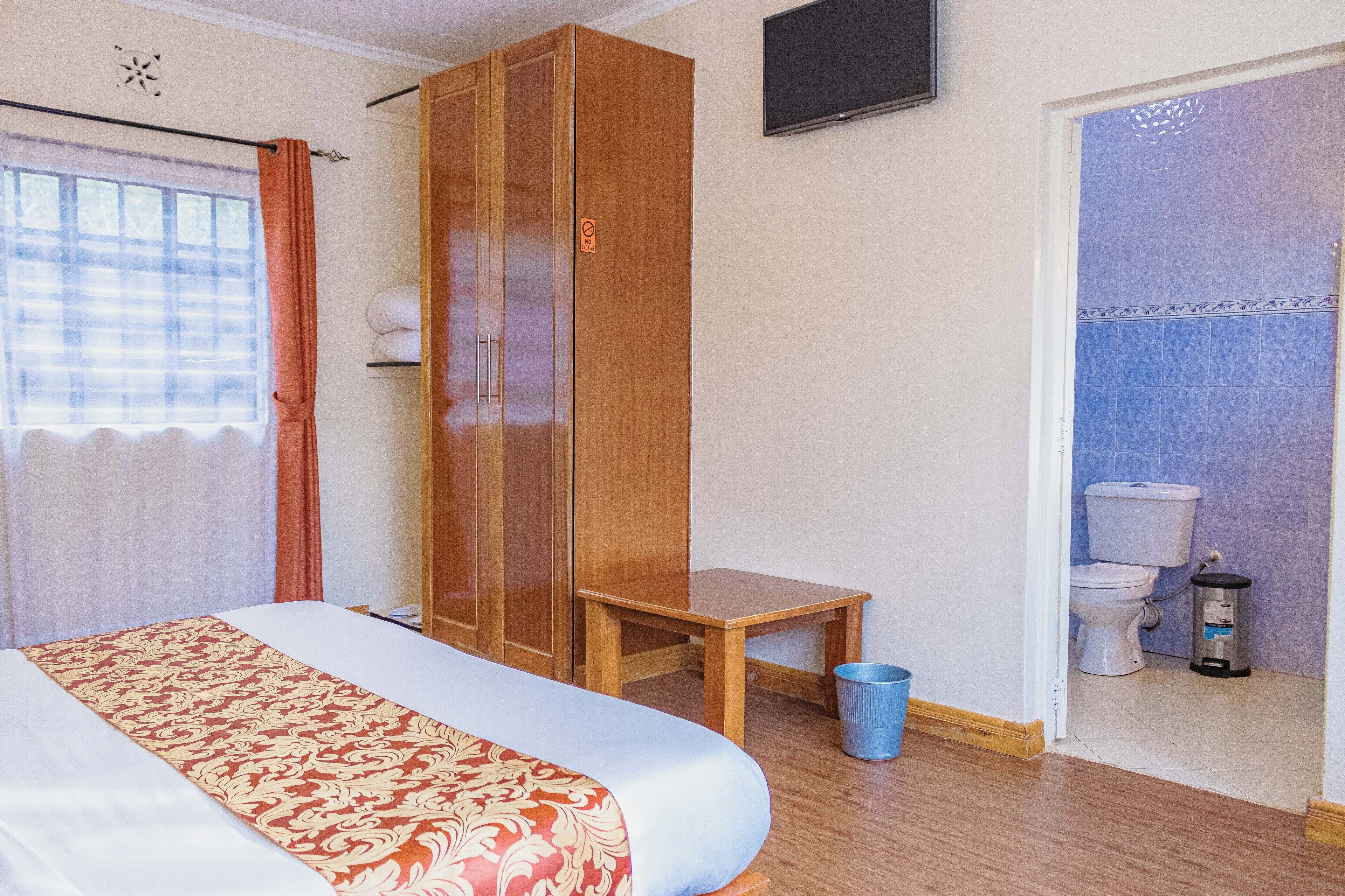 Quarto de hotel com uma cama, um guarda-roupa de madeira, uma mesa pequena, um cesto de lixo e um banheiro com vaso sanitário e pia.