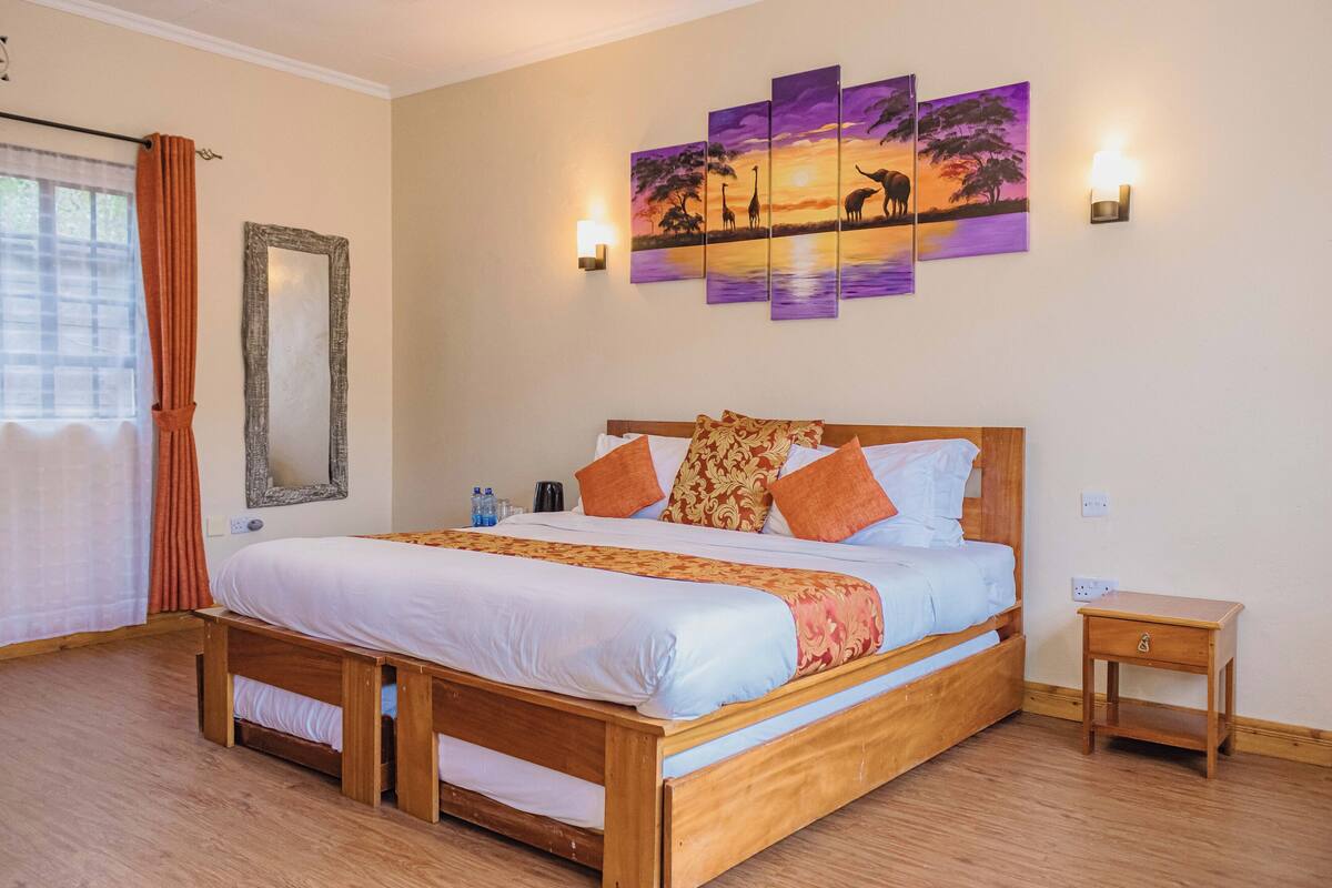 A bedroom with a wooden bed, orange and white bedding, a wall-mounted mirror, a sunset painting, and a bedside table.